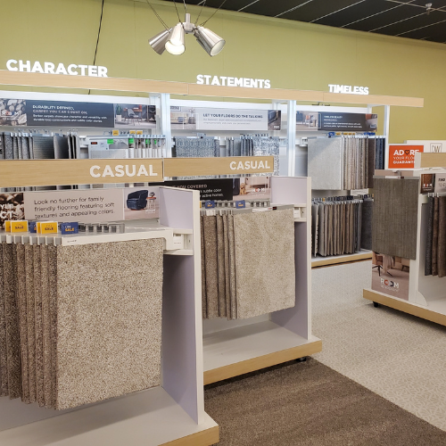 Interior of The Floor Factory showroom with wall displays of carpet options with “CHARACTER,” “STATEMENTS,” and “TIMELESS,” plus rows of carpet samples in “CASUAL” sections and a hanging ceiling light.